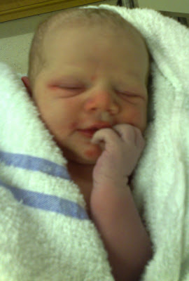 Flashback Friday: Newborn baby wrapped in a white towel with blue stripes, lying down with eyes closed and one hand near their mouth, appearing peaceful and content.