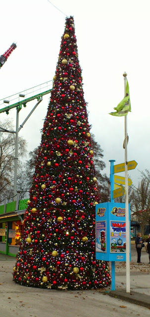 A tall, outdoor Christmas tree adorned with red, gold, and white ornaments stands beside a signpost and flag at an amusement park on a cloudy day—perfect for a Flashback Friday memory.