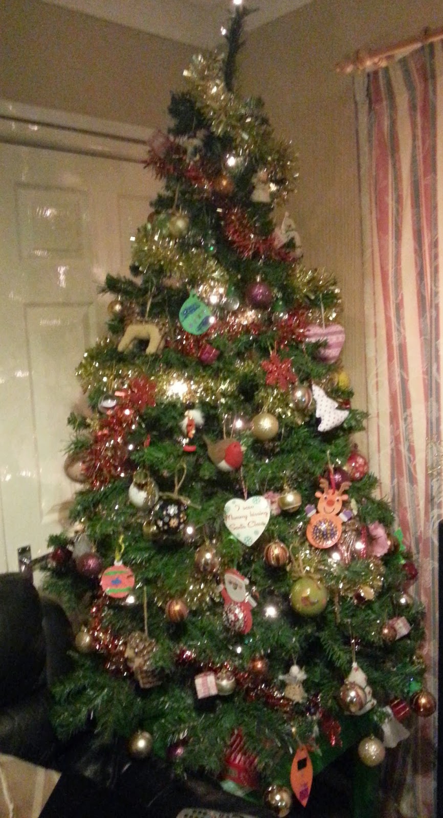 A decorated Christmas tree stands indoors, adorned with colourful baubles, tinsel, fairy lights, and handmade paper decorations. Wrapped presents—some meant to save money and others the most expensive—are placed underneath the tree.