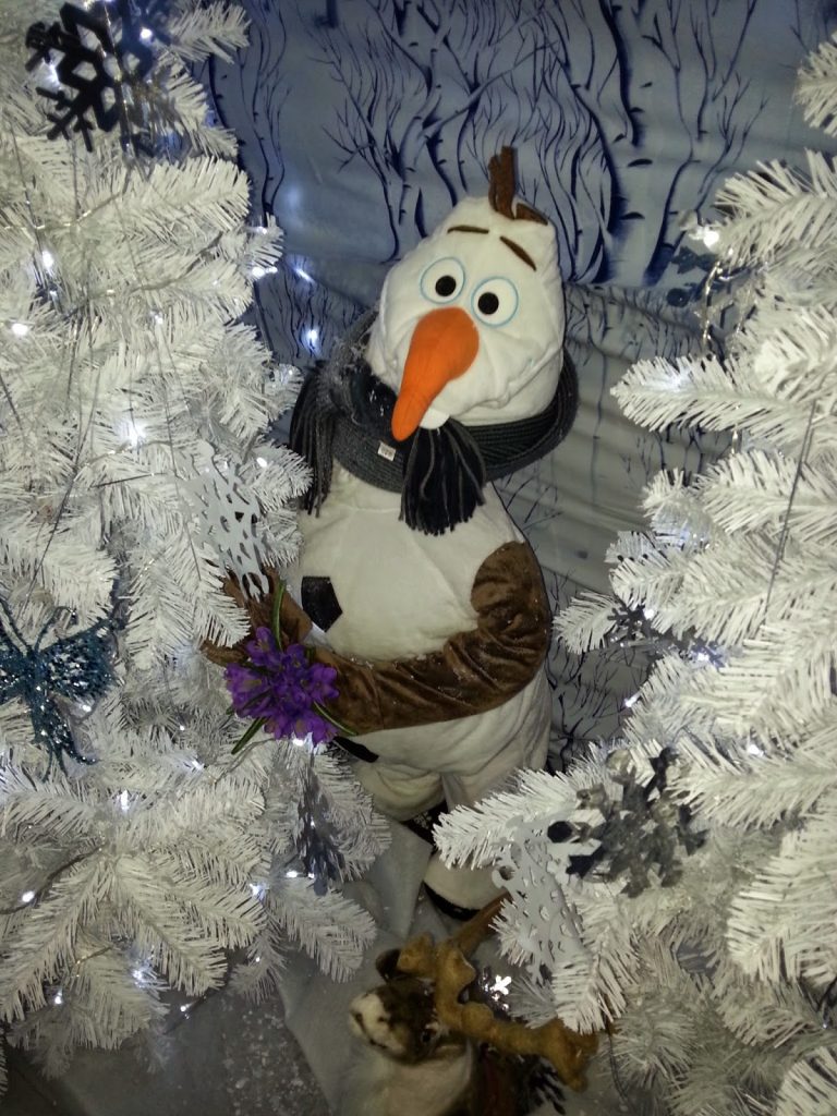 A plush Olaf snowman from Frozen stands among white artificial Christmas trees with snowflake ornaments, holding a purple flower, basking in glowing white lights and a snowy forest backdrop—a perfect Silent Sunday moment.