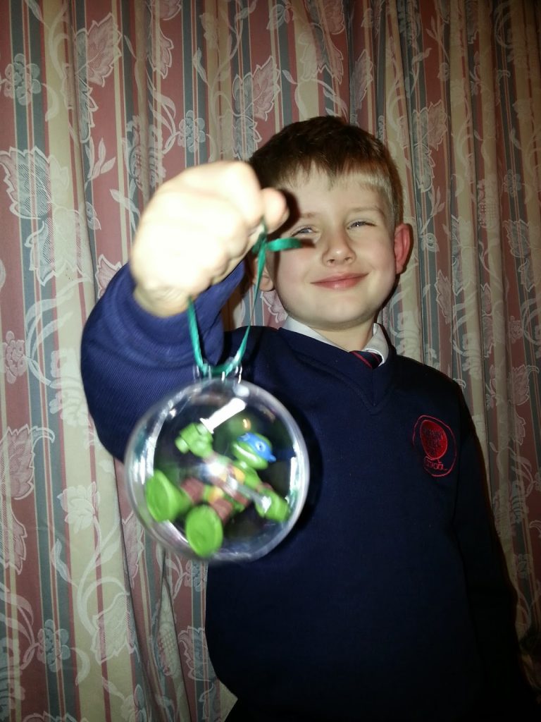 James, a young boy in a blue school uniform, smiles as he holds up a clear bauble with a small green toy inside, in front of patterned curtains. Happy Christmas!.