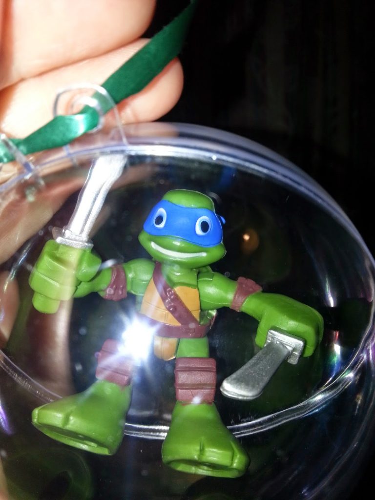 A hand holds a clear plastic bauble containing a small Teenage Mutant Ninja Turtle with a blue mask, holding a silver sword—ready for action. Happy Christmas from James!.