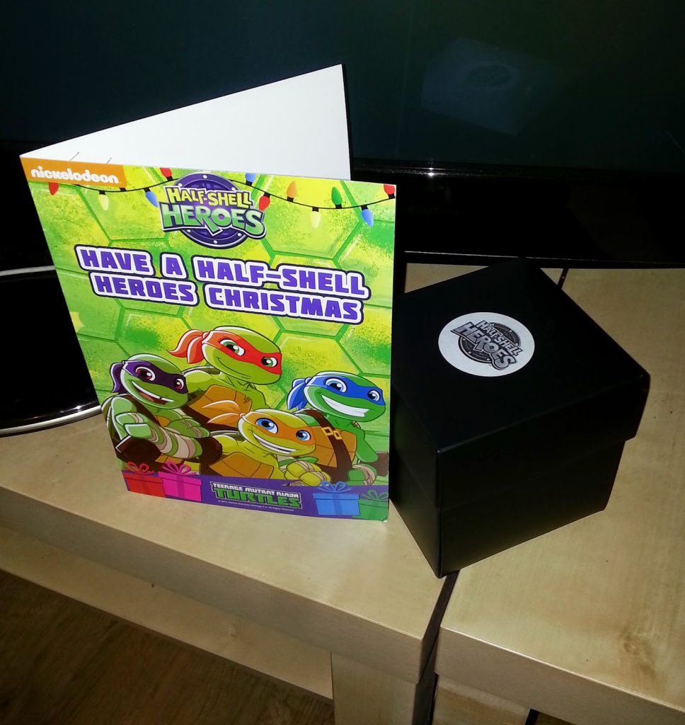 A Teenage Mutant Ninja Turtles Christmas card stands upright next to a closed black gift box on a wooden table in front of a TV. The card, perfect for James, features the turtles in Santa hats and reads, "Have a Half-Shell Heroes Merry Christmas.