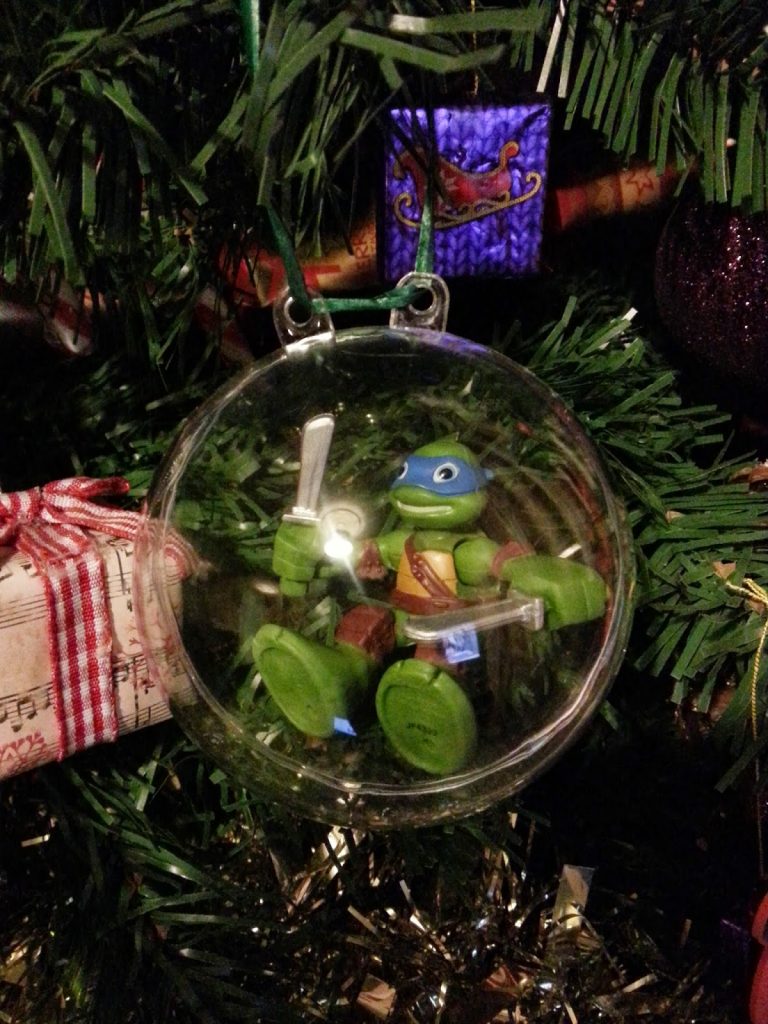 A Teenage Mutant Ninja Turtle figurine holding two swords is enclosed in a clear bauble, hanging on a decorated Christmas tree with green branches and decorations. Merry Christmas, James—a perfect festive touch by the wrapped present.