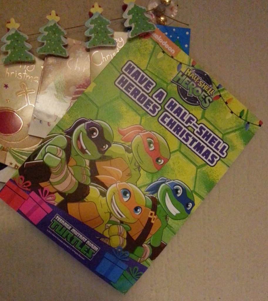 A Teenage Mutant Ninja Turtles Christmas card with “Have a Half-Shell Heroes Christmas” text is placed on a table, surrounded by gift tags decorated with Christmas trees and a handwritten Merry Christmas from James.
