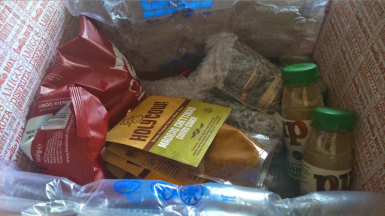 A snack subscription box filled with various packaged snacks, including crisps, nut butter sachets, two small beverage bottles, and an item wrapped in bubble wrap, all surrounded by air-filled packing cushions.