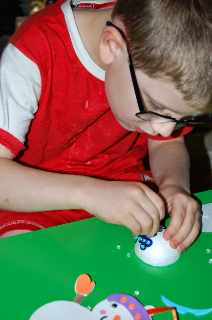 A young boy wearing glasses and a red shirt carefully decorates a white foam ball with small blue pieces, making holiday savings crafts while sitting at a green table with supplies scattered around.