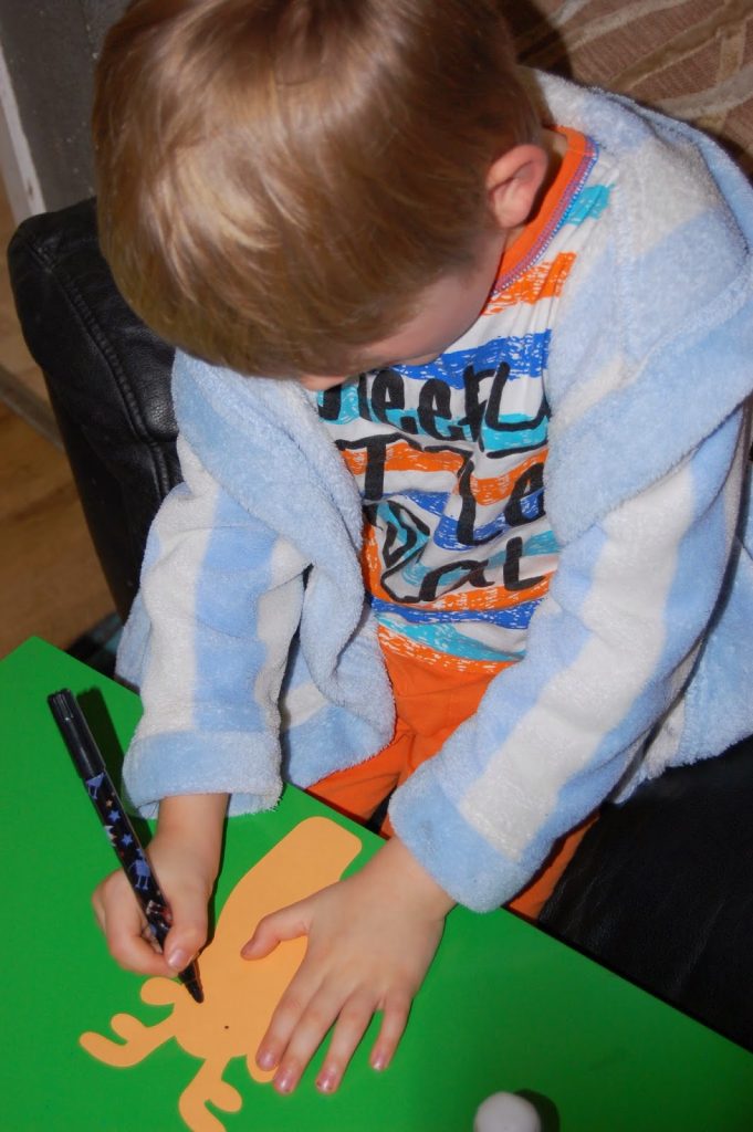 A young child wearing a striped shirt and blue dressing gown draws on an inexpensive yellow paper cut-out of a reindeer placed on a green surface, using a black marker pen.