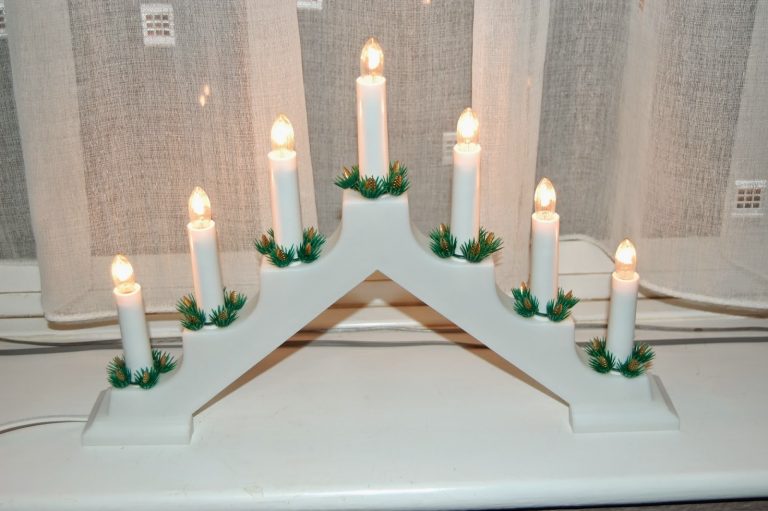 A white, electric candelabra with seven lit candle-shaped bulbs in a V-shape, trimmed with small green artificial greenery at each base, sits on a windowsill before sheer curtains—perfect for decking the halls and adding festive charm to your home décor.
