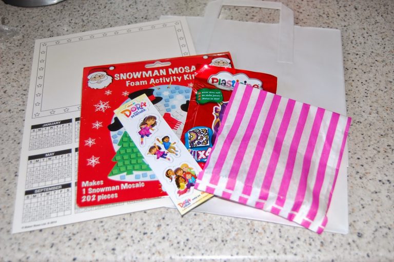 A fun craft party set featuring a snowman mosaic kit, plasticine, Dora the Explorer Nick Jr stickers, a striped pink-and-white bag, a white gift bag, and activity sheets displayed on a speckled worktop.