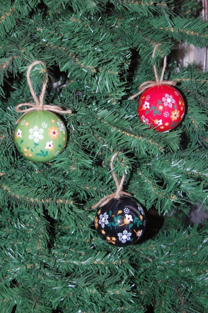 Three decorated Christmas baubles—green, red, and black with painted flowers—hang on a green artificial Christmas tree, each tied with string. Perfect for decking the halls and adding festive charm to your holiday decorations.