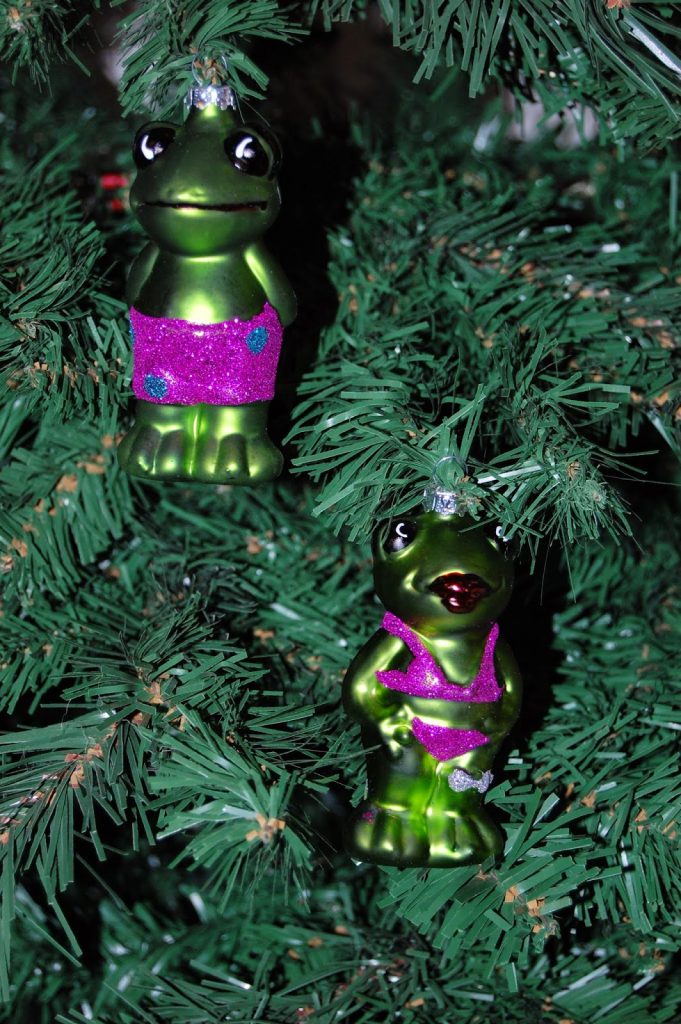 Two green frog ornaments in sparkly pink outfits hang on the Christmas tree, their large eyes and shiny details adding a playful touch to your holiday decorating and home décor.