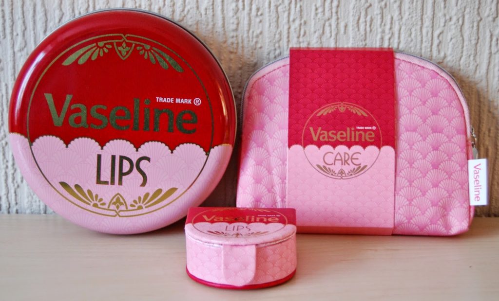 A large red and pink Vaseline Lips tin, a matching small tin, a pink Vaseline Care zippered pouch, and a small rectangular box are displayed against a textured wall—perfect gifts for her.