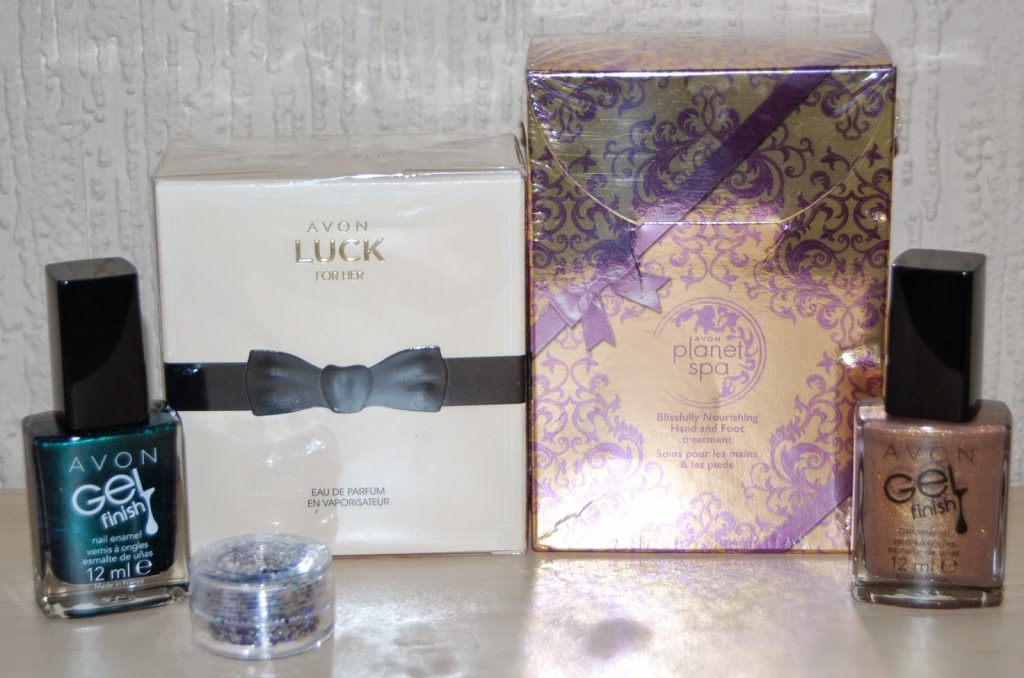 Two Avon Gel Finish nail polishes, a small glitter pot, Avon Luck for Her perfume in a white box with a bow, and Planet Spa Blissfully Nourishing hand and foot cream make perfect gifts for her, beautifully displayed on a table.
