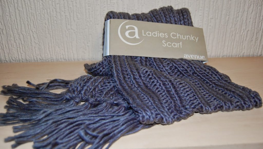 A folded, dark grey chunky knit scarf with fringed ends sits on a light wooden surface. Its label reads "Ladies Chunky Scarf" and "avenue." in white text on beige—an ideal choice among cosy gifts for her.