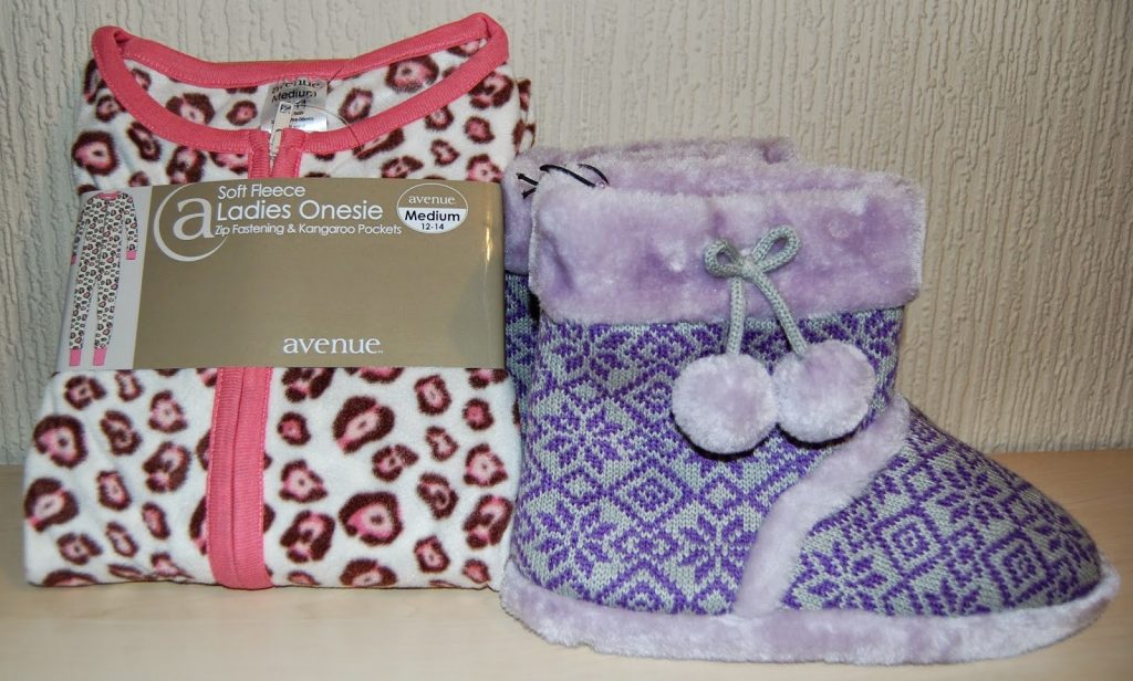 A folded white and pink leopard print fleece onesie with a label sits next to purple patterned slipper boots with pom-poms—perfect gifts for her—displayed on a light surface against a textured wall.