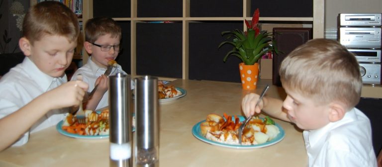 Three young boys in white shirts sit at a dining table eating a meal with vegetables and meat. A plant in an orange pot and a stereo are visible in the background, creating a cosy family dinner atmosphere as the boys focus on their food.