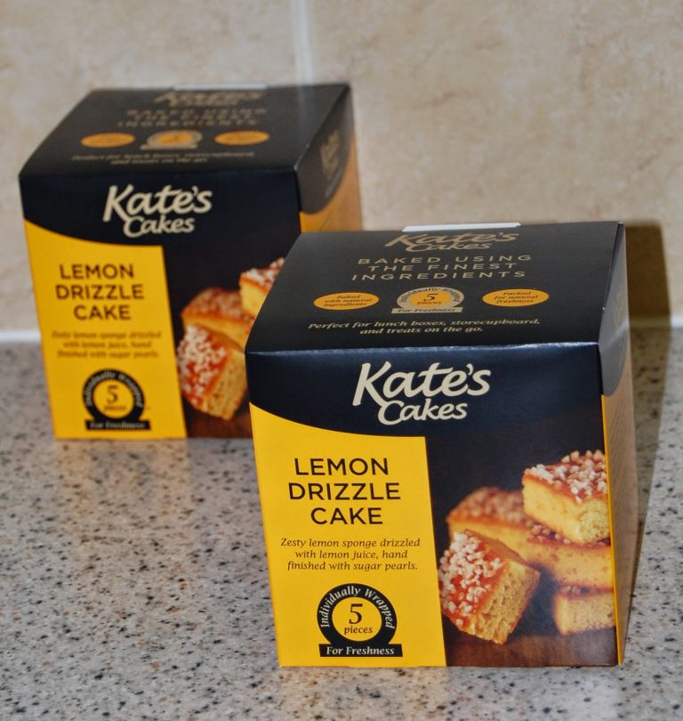 Two boxes of Kate's Cakes Lemon Drizzle Cake sit on a kitchen worktop. The black and yellow boxes show images of sliced lemon drizzle cake with sugar pearl toppings—perfect to enjoy with your favourite brew.