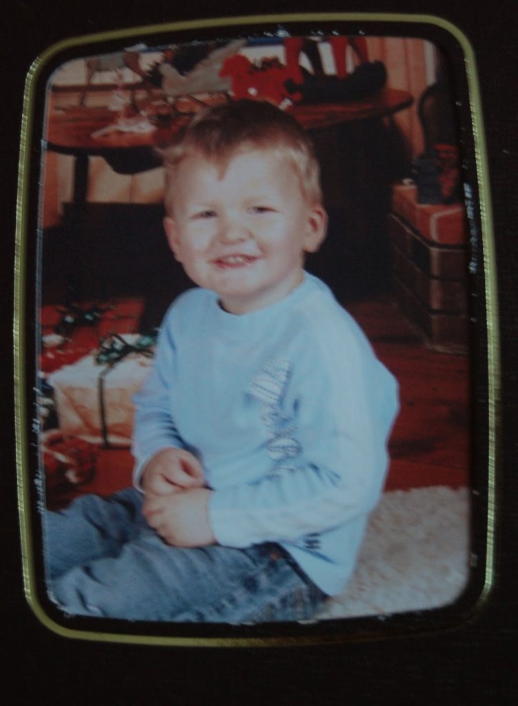 Flashback Friday: A young child with short blonde hair, wearing a light blue shirt and jeans, sits smiling on a rug indoors with wrapped presents and holiday decorations in the background.