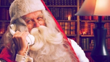 Father Christmas, wearing a red hat and spectacles, smiles whilst talking on a white rotary telephone. Seated in a cosy room lined with books, he shares Personalised Christmas Messages by the warm glow of a lamp, bringing joy from the Portable North Pole.