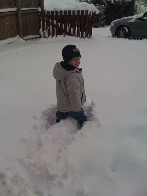 Flashback Friday: A young child in a winter coat, hat, and gloves stands outside in deep snow, with snow reaching above their knees. A wooden fence and a car are visible in the background.