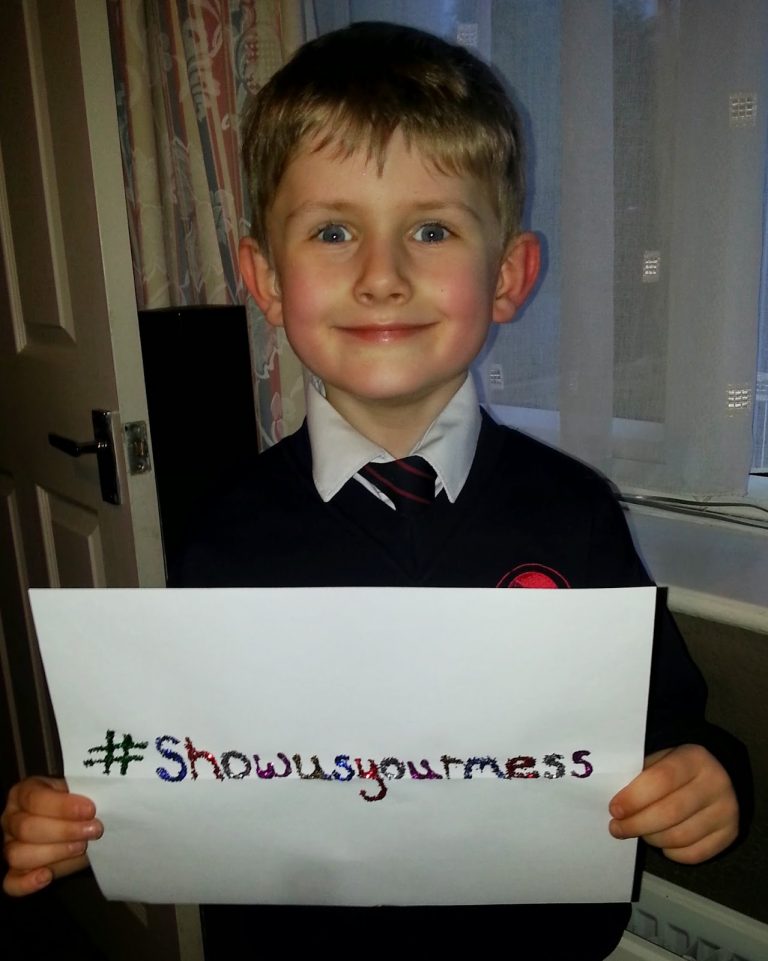 A young boy in a school uniform smiles whilst holding a handwritten Bosch sign that says "#Showusyourmess." He stands indoors in front of a window with curtains.