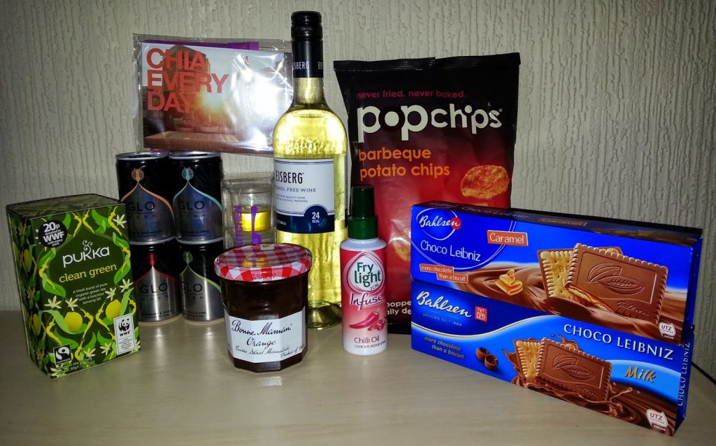 A curated snack subscription spread sits on a table—tea, energy drinks, wine, crisps, chocolate biscuits, chia seeds, jam, and chilli oil spray—perfect for a Degustabox review against a textured wall backdrop.