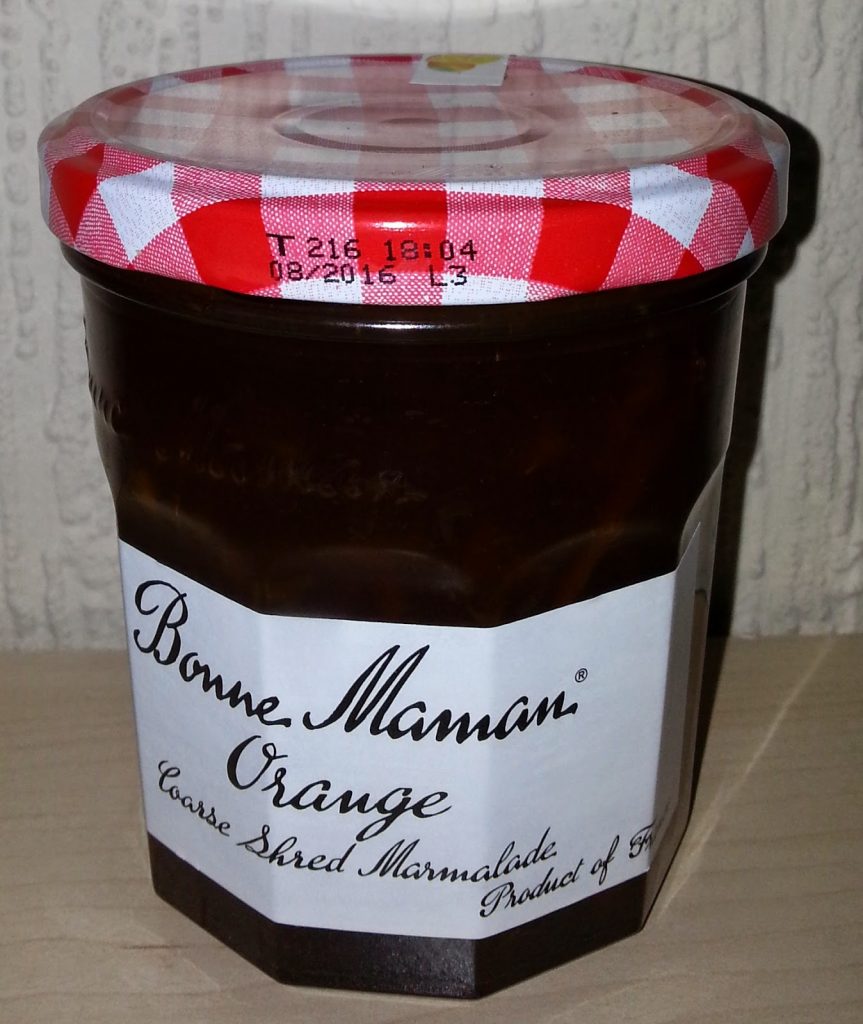 A jar of Bonne Maman Orange Coarse Shred Marmalade with a red and white chequered lid, featured in a Degustabox subscription box, sits on a wooden surface in front of a textured white wall.