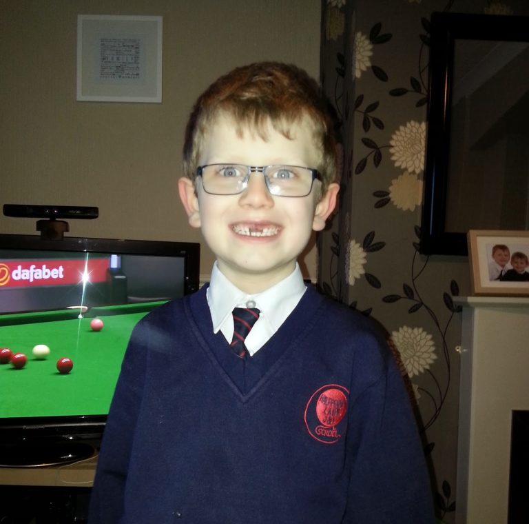 A young boy with glasses smiles at the camera, wearing a blue school uniform and striped tie. It’s a true Silent Sunday scene, with a snooker match on the telly and family photos arranged on the white unit behind him.