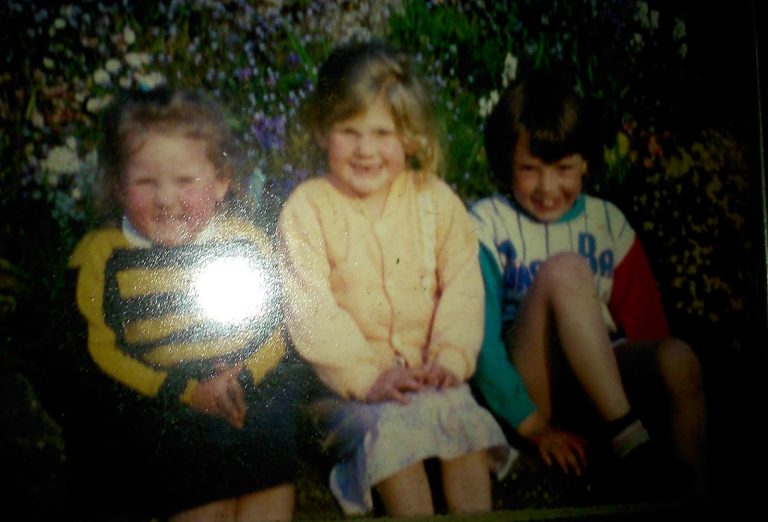 Three young children sit on the ground outdoors, smiling at the camera for a perfect Flashback Friday moment. The child on the left wears a yellow and black striped jumper, while the others sport cosy cardigans and stripes. A slight glare adds to the nostalgic feel.