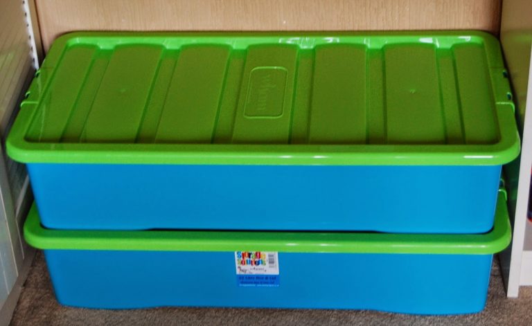 Two large rectangular plastic storage boxes with bright green lids and blue bases are stacked on a carpeted floor under a wooden shelf, offering an ideal toy organisation solution to neatly store toys out of sight.