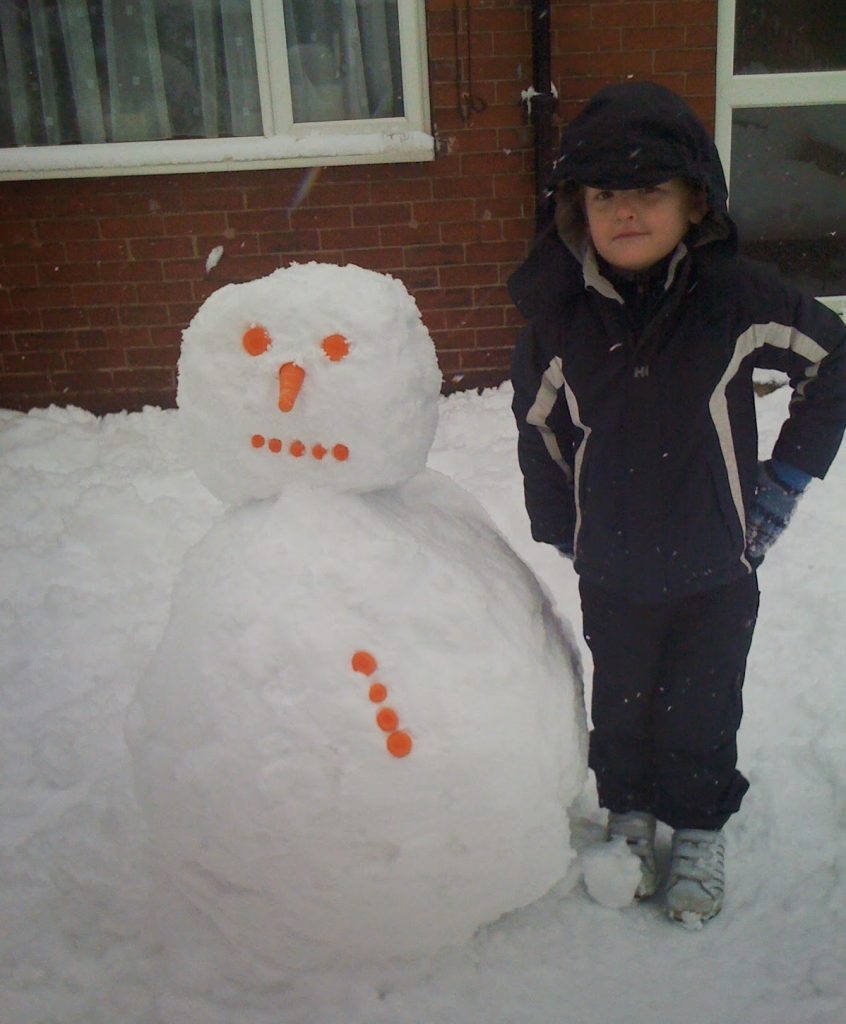 Flashback Friday: A child in a hooded winter coat stands next to a snowman with orange buttons for eyes, mouth, nose, and decorations, in a snowy garden in front of a brick house.