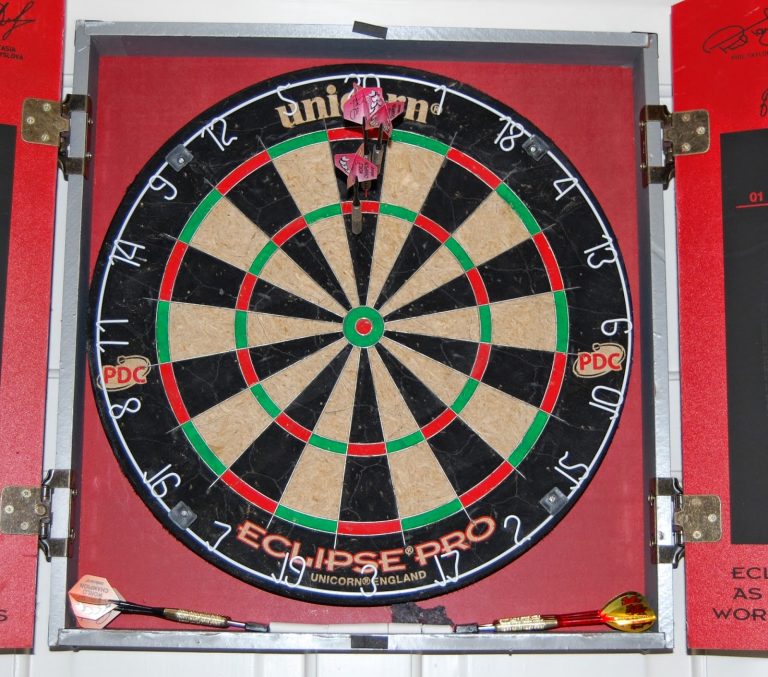 A dartboard with three darts clustered in the bullseye and two in the tray below, set in a red cupboard with visible hinges—capturing a perfect Silent Sunday moment, with partial writing on the doors adding to the scene.