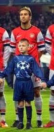 Flashback Friday: A footballer in an Arsenal shirt stands behind a young boy in a blue kit, holding hands with someone out of frame before a match.