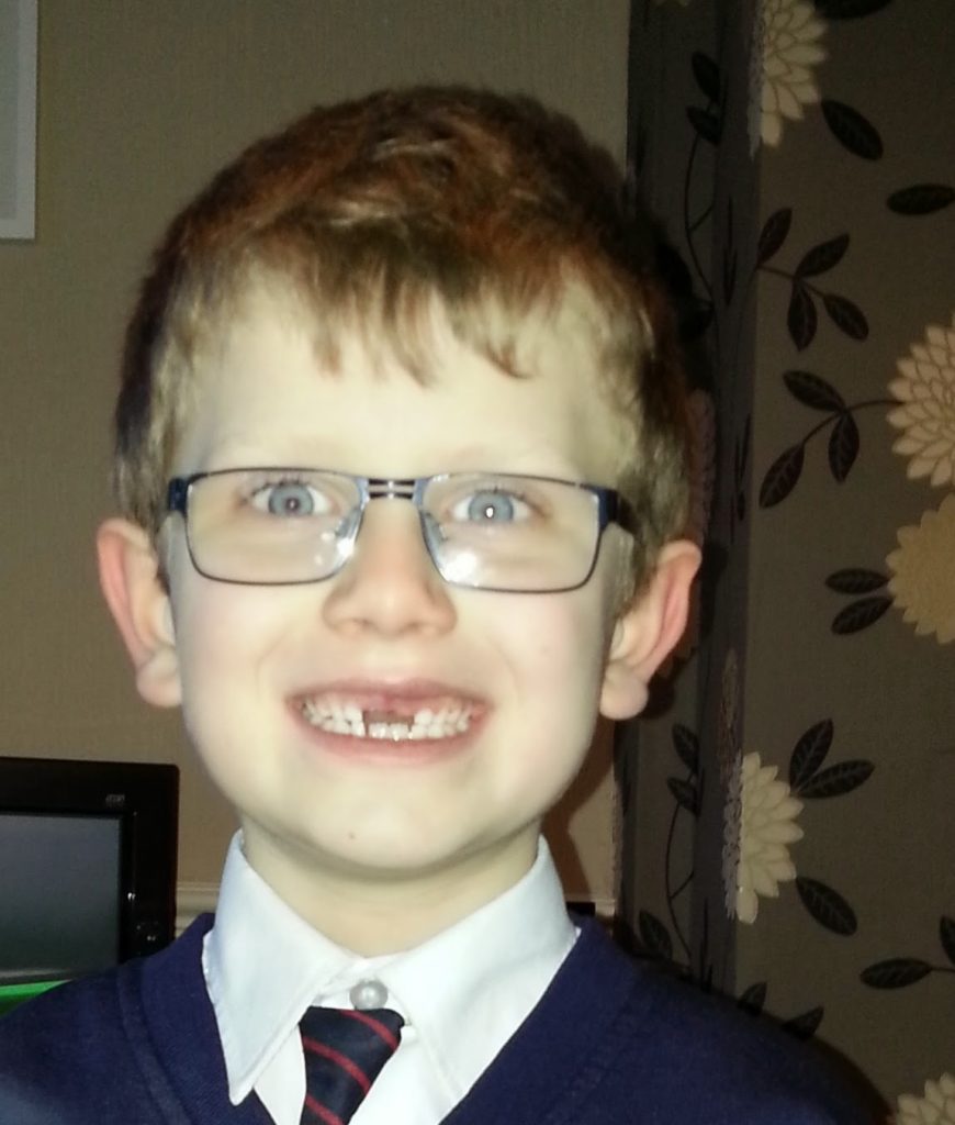 A young boy with glasses and short light brown hair smiles, showing missing front teeth. Smartly dressed, he’s ready for a lesson on the value of money. The background includes a floral-patterned wall and part of a TV screen.
