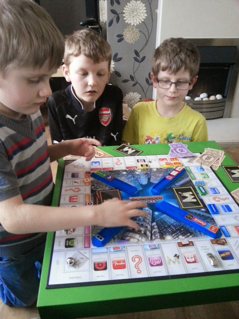 Three young boys sit around a table for a Silent Sunday, playing Monopoly. One boy moves a piece on the board whilst the others watch attentively, with Monopoly money and cards scattered across the table.