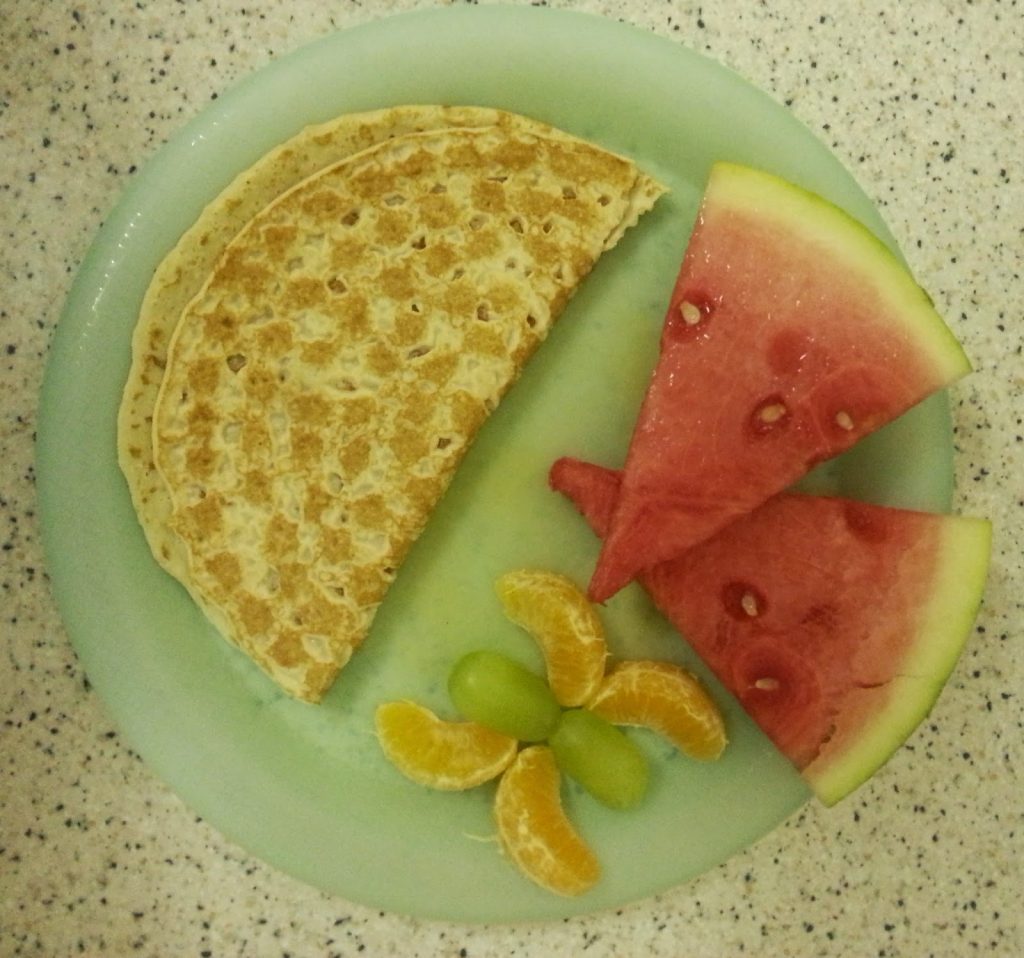 A plate with half an Abra-ca-Debora pancake, two watermelon slices, orange segments, and green grapes arranged in a decorative pattern on a speckled worktop—perfect for Pancake Day.