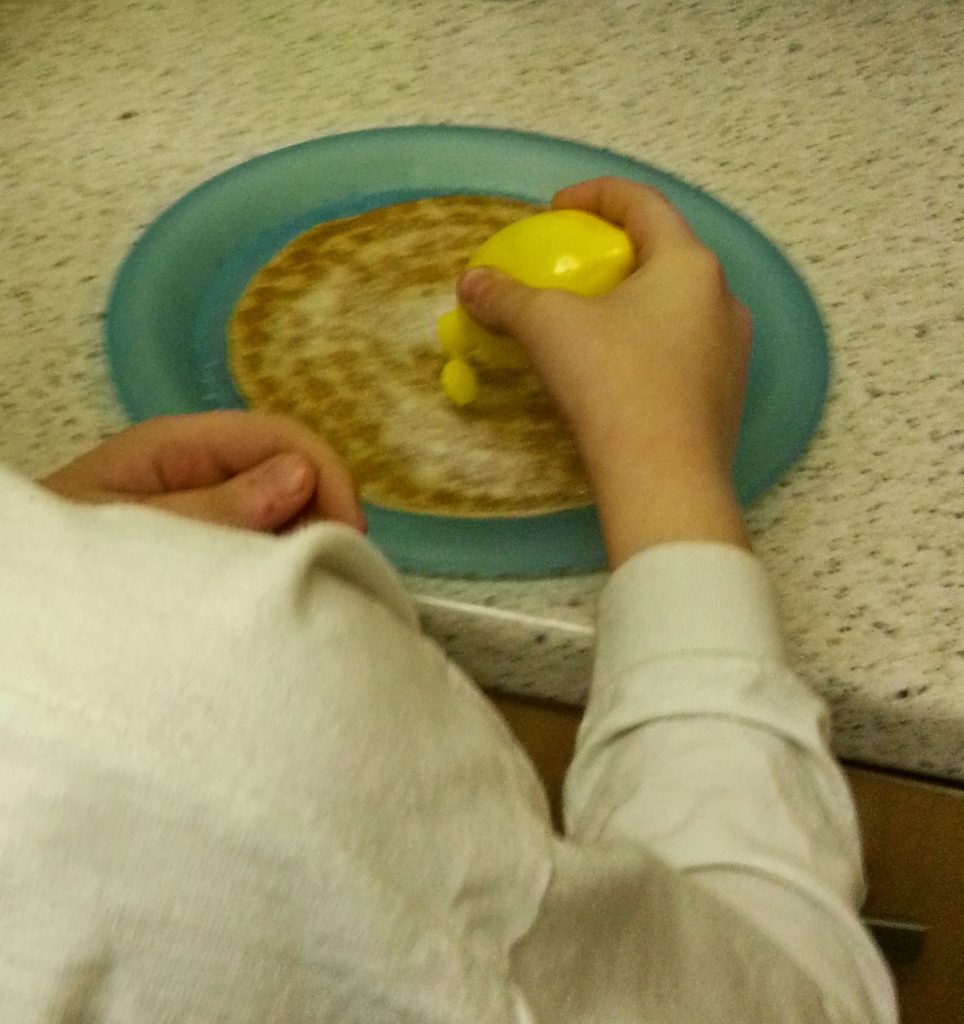 A person squeezes a yellow lemon-shaped bottle over a pancake on a blue plate, celebrating Pancake Day with an Abra-ca-Debora treat on the worktop.
