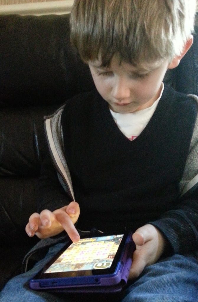 A young boy is sitting on a dark sofa, focused on playing a game on his Kindle Fire HD 6. He uses his index finger to touch the screen, and the tablet is in a purple case, making it perfect for any tablet review.