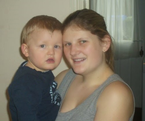 A young woman with long hair smiles whilst holding a toddler with short hair. They are indoors, standing close together in front of a window with sheer curtains—such a sweet Flashback Friday moment.