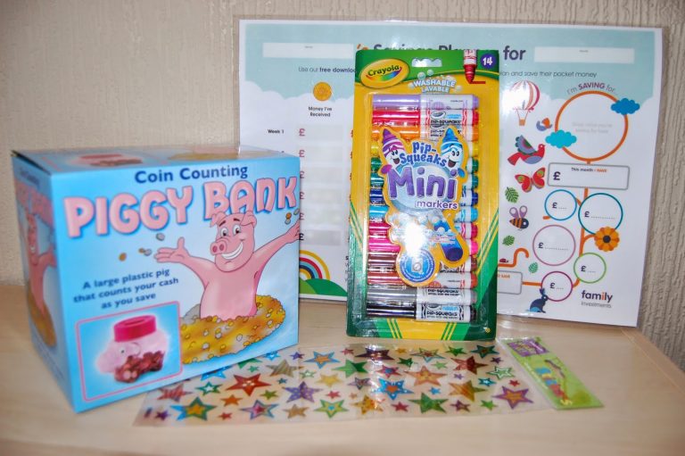 A box labelled "Coin Counting Piggy Bank," a pack of Crayola Pip-Squeaks Mini Markers, colourful star stickers, and a savings goals chart are displayed on a wooden surface, perfect for teaching children about money and the value of money.