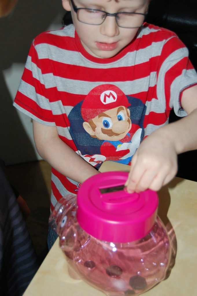 A child wearing glasses and a red-and-white striped Mario shirt is putting coins into a pink piggy bank, teaching children about money and helping them learn the value of money from an early age.