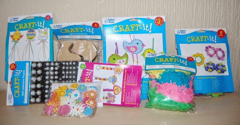 A variety of colourful Baker Ross Spring Crafts Craft-it! kits with materials such as foam masks, beads, embellishments, and shredded tissue, all displayed on a wooden surface against a beige wall.