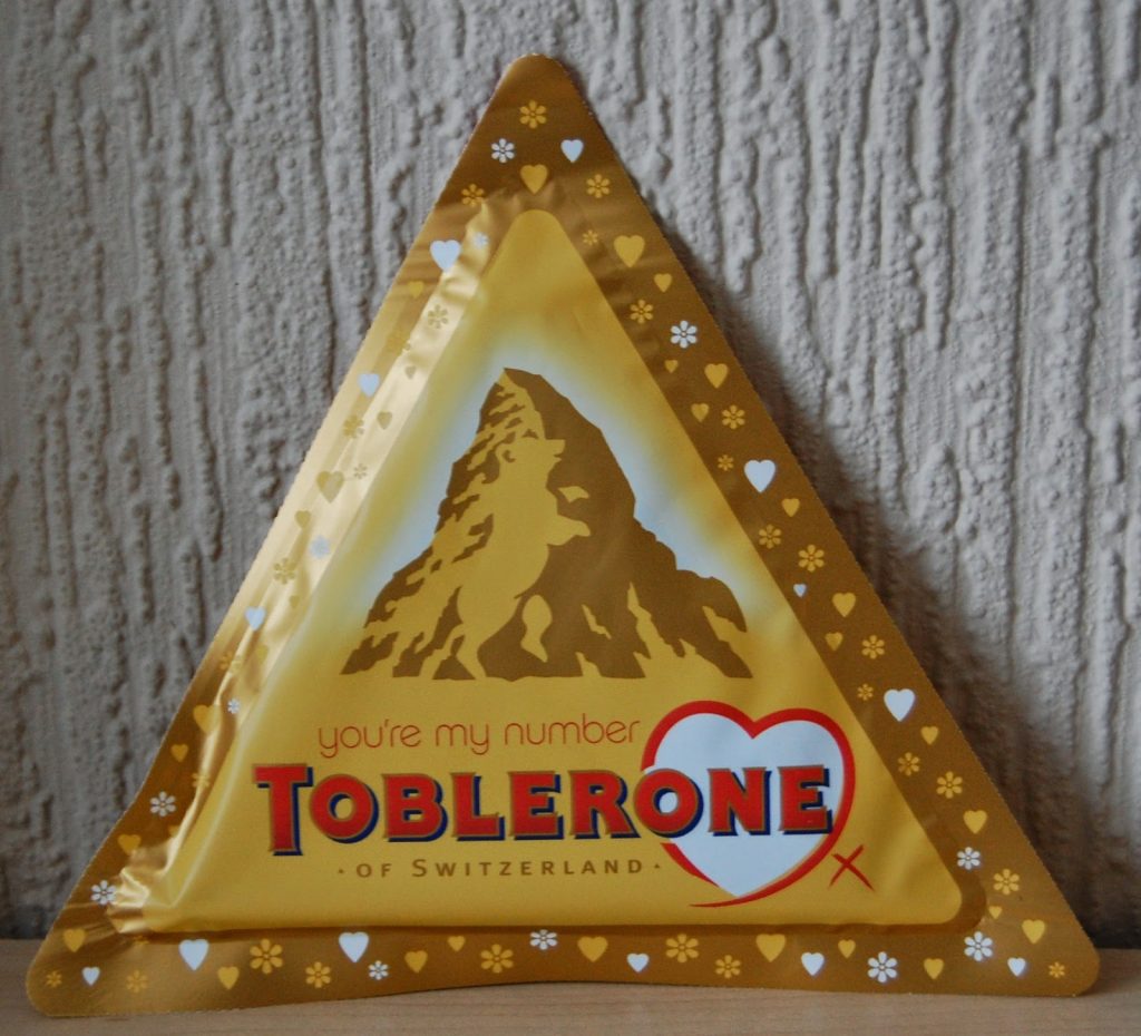 A triangular yellow Toblerone chocolate tin with a mountain illustration and the words "you're my number Toblerone" in red and orange, set against a textured beige wall—perfect for Valentine’s Day gift ideas.
