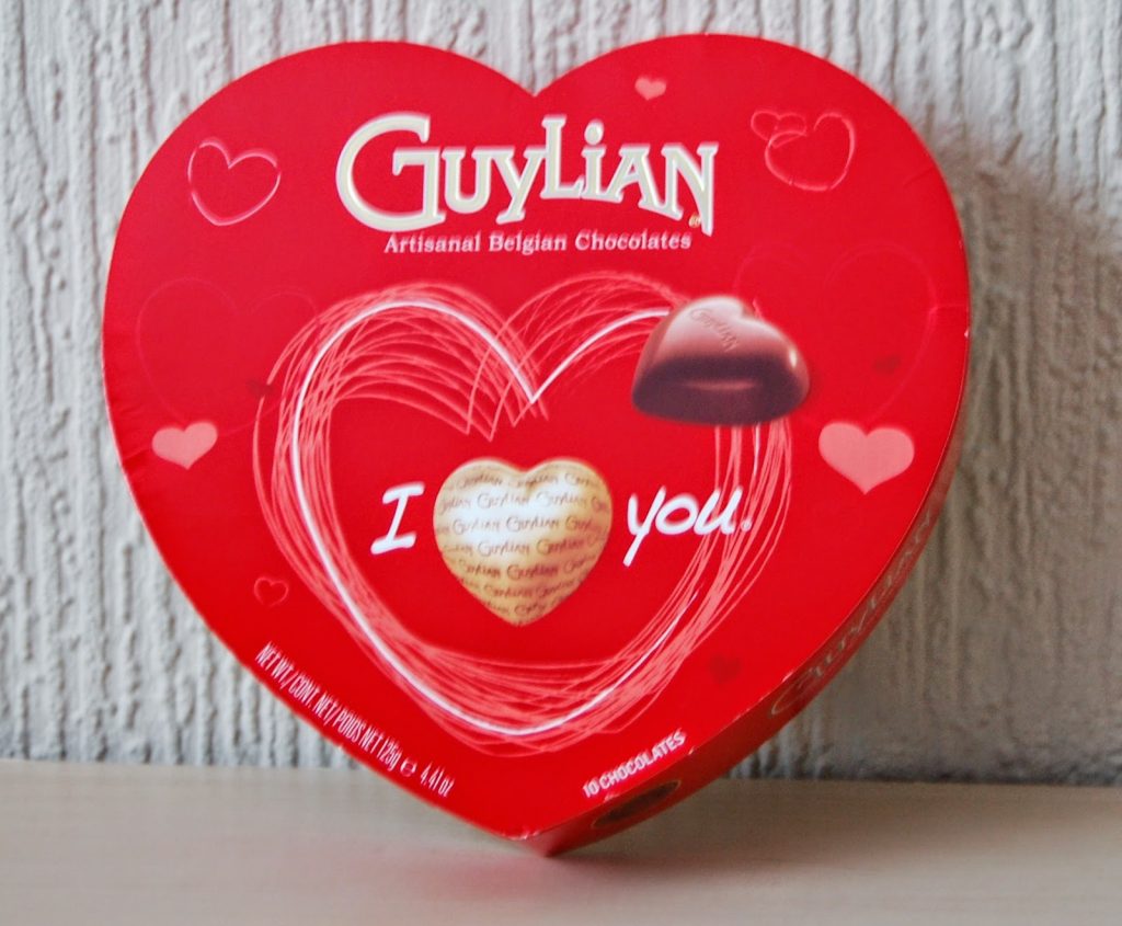 A red, heart-shaped box of Guylian Belgian chocolates with white and red heart designs and "I love you" on the lid—an ideal choice for Valentine’s Day gift ideas—sits in front of a textured white wall.