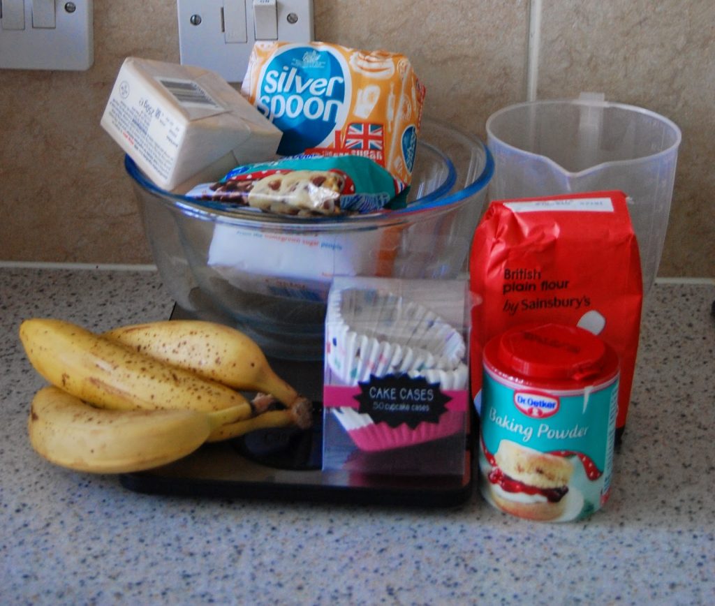 A collection of baking ingredients on a kitchen worktop, ready to make Banana Chocolate Chip Muffins, including bananas, flour, baking powder, cake cases, butter, sugar, eggs, and a clear measuring jug.