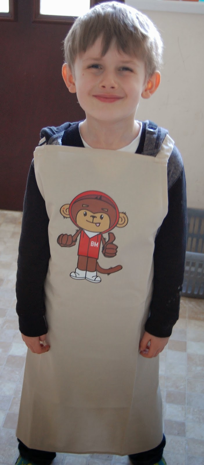A young boy stands indoors, smiling and wearing a beige apron with a cartoon monkey mascot dressed in red on the front—perfect for baking banana muffins or chocolate chip treats. The background shows a tiled floor and a dark door.