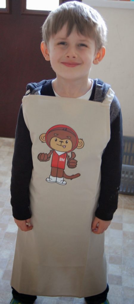 A young boy stands indoors smiling, wearing a tan apron featuring a cartoon monkey in a red outfit with a helmet labelled "BM," ready to bake Banana Chocolate Chip Muffins. The background includes a door and a plastic basket.
