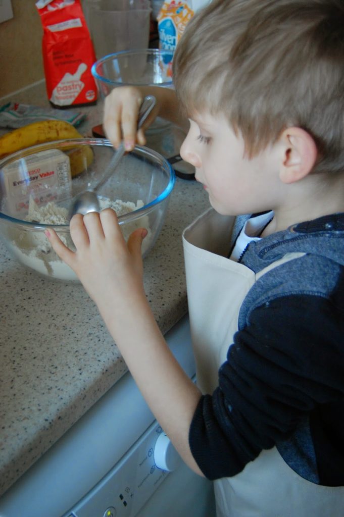 A young child wearing an apron mixes ingredients in a glass bowl on a kitchen worktop, preparing a banana muffins recipe. Baking supplies, including flour and bananas, are visible in the background.