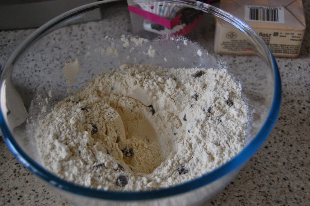 A glass bowl on a speckled worktop contains flour mixed with sultanas for Banana Chocolate Chip Muffins, with a well in the centre ready for wet ingredients. Boxes and muffin cases are visible in the background.